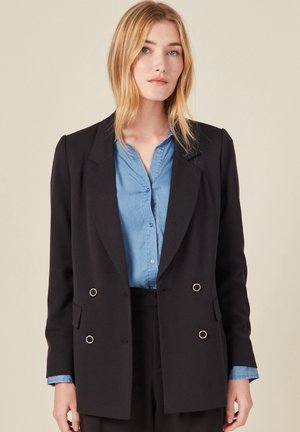 Blazer negro de doble botonadura con botones de tono dorado, solapas con muesca y un ajuste entallado, superpuesto a una camisa de chambray azul.