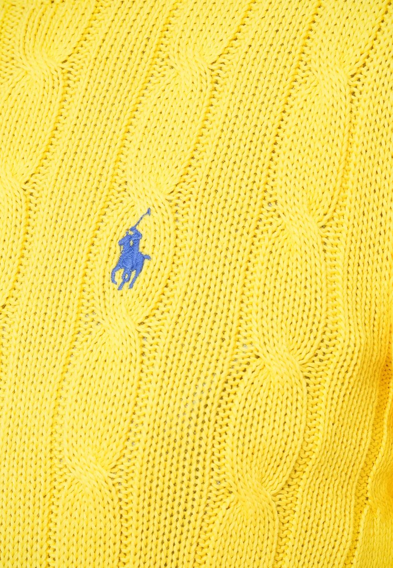 Gele gebreide trui met een kabelpatroon en een klein blauw geborduurd logo. De textuur is zacht met details van open steken.