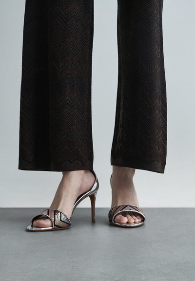 Σανδάλια με ψηλό τακούνι που διαθέτουν μια μεταλλική ασημί λωρίδα στη μύτη και υφασμάτινη επένδυση με σχέδια, συνδυασμένα με έναν κομψό, γυαλιστερό τακούνι και ανοιχτή μύτη.