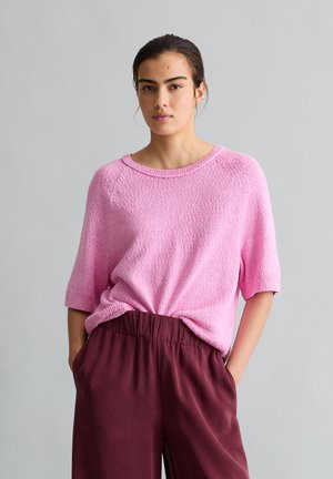 Vrouw staat voor een grijze achtergrond, draagt een gestructureerde roze korte mouwen trui en bordeaux elastische taillebroek, handen in de zakken.