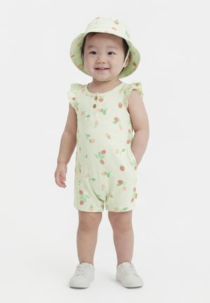 Tout-petit debout, portant une combinaison légère verte à imprimé fraises, un chapeau assorti et des baskets blanches, souriant sur fond blanc.