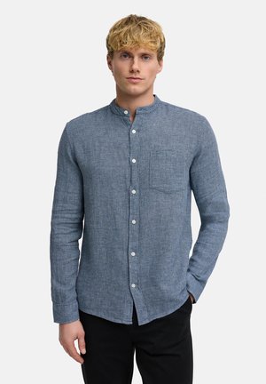 Nuori mies, jolla on vaaleat hiukset, pukeutuneena siniseen teksturoituun nappipaitaan mandariinikauluksella ja mustiin housuihin, seisoo käsi taskussa.