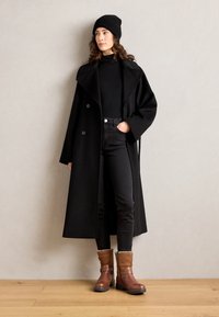 Langer schwarzer Wollmantel mit großen Revers, kombiniert mit einem schwarzen Rollkragenpullover, schwarzen Skinny Jeans und braunen Stiefeletten mit Fellfutter.