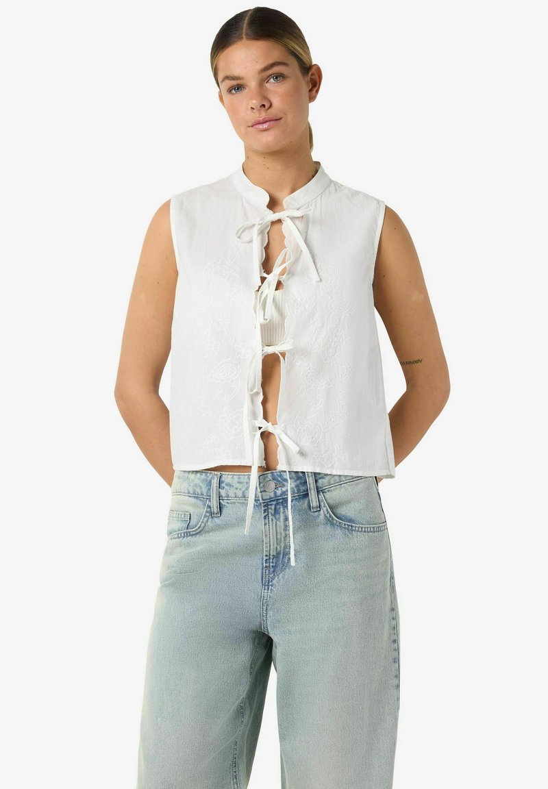 Femme portant un haut blanc sans manches avec des liens à l'avant et un jean bleu clair taille haute, debout les mains derrière le dos.