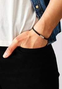 Bracciale in pelle intrecciata nera con un accento metallico argento, indossato su un polso. Lo sfondo mostra una mano appoggiata in una tasca.