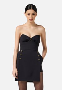 Donna con capelli scuri in un mini abito nero senza spalline con bottoni dorati sulle tasche anteriori, in piedi contro uno sfondo chiaro e uniforme.