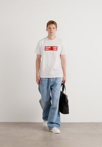 Balts īsām piedurknēm T-krekls ar sarkano "Calvin Klein" logotipu, gaiši zili brīvi pieguļoši džinsi un balti sporta apavi. Melna soma ar rokturiem.