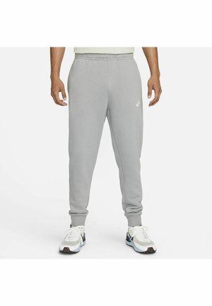 Homme portant un pantalon de jogging Nike gris clair et des baskets blanches, debout les bras détendus le long du corps devant un fond uni.