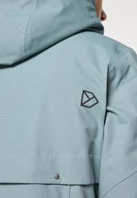 Jasnoniebieska wodoodporna kurtka z kapturem, teksturowanym materiałem i geometrycznym czarnym logo na lewym ramieniu. Srebrne detale z zatrzaskami.