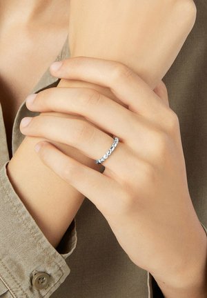 Hand mit einem silbernen Perlenring am Ringfinger, Finger halten sanft das gegenüberliegende Handgelenk, beiges Hemd sichtbar.