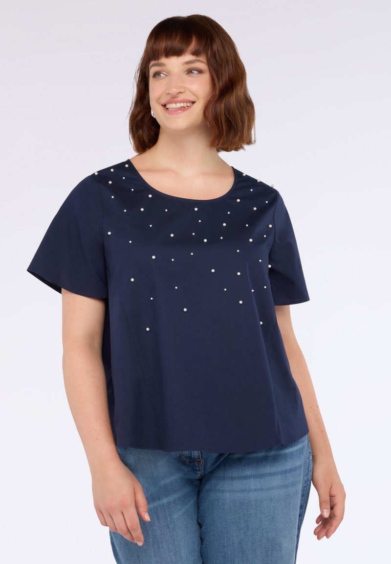 Donna sorridente, che indossa una blusa a maniche corte blu navy con piccole perle bianche sparse e jeans blu, in piedi contro uno sfondo bianco.