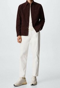 Camicia marrone con bottoni e tasca sul petto, abbinata a jeans tapered avorio chiaro e sneakers beige chiare. Texture liscia su tutto.