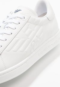 Sneaker in pelle bianca con dettagli testurizzati, lacci piatti e suola in gomma con motivo. Presenta un marchio in rilievo sul lato e sulla linguetta.