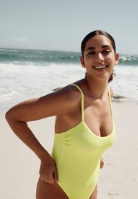 Neoninės geltonos spalvos vientisas maudymosi kostiumėlis su tekstūruota medžiaga, plonais dirželiais ir žemu iškirptės iškirpimu, fonas – paplūdimys su jūros banga.