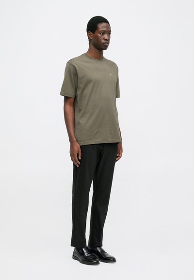 T-shirt a maniche corte di colore verde oliva con collo rotondo, indossata con pantaloni neri e scarpe nere, caratterizzata da un design minimalista senza motivi.