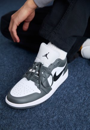 Personne portant des baskets Nike Air Jordan blanches et grises avec un swoosh noir, des chaussettes blanches et un pantalon noir, assise sur un tapis bleu.