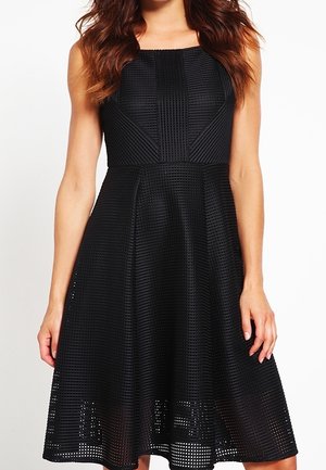 Femme portant une robe noire texturée sans manches en ligne A avec un décolleté carré et un ourlet à hauteur des genoux.