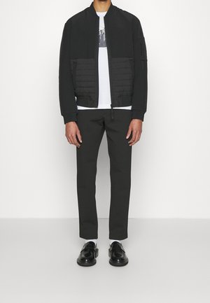 Svart bomberjacka med vadderad nedre sektion, kombinerad med mörka byxor och svarta skor. Modellen bär en vit t-shirt under.