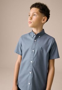 Vaaleansininen, lyhythihainen nappipaita, jossa on sileä pintakudos, pieni kirjailtu logo rinnassa ja valkoiset napit.