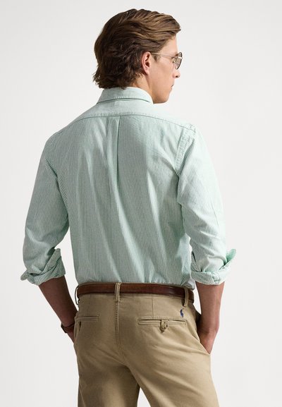 Green and white striped button-up shirt with rolled sleeves, paired with tan pants featuring back pockets and embroidered logo on the right.