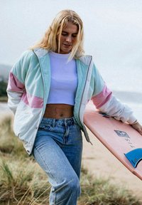 Giovane donna con capelli biondi che indossa una giacca pastello e jeans, porta una tavola da surf rosa su una spiaggia erbosa.