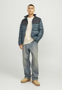 Giacca imbottita di un blu scuro con colletto nero, abbinata a un maglione beige chiaro, jeans larghi e stivali beige. Tessuto morbido, vestibilità casual.