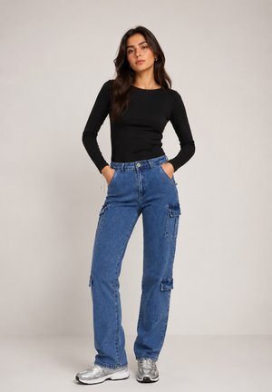 Frau steht mit den Händen in den Taschen, trägt ein schwarzes Langarmshirt, blaue Cargo-Jeans und silberne Turnschuhe vor einfachem Hintergrund.