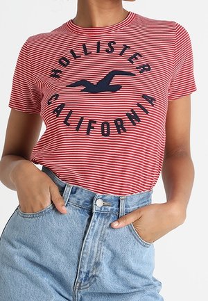 Osoba nosząca czerwoną i białą koszulkę Hollister California w paski, wpuszczoną w jasnoniebieskie, wysokie jeansy, ręce w kieszeniach.