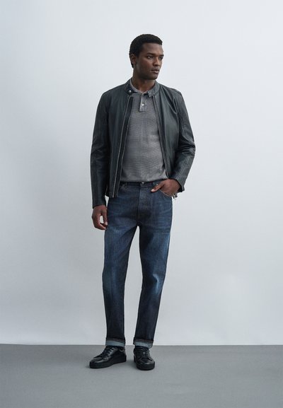 Άνδρας με μαύρο δερμάτινο μπουφάν, γκρι πόλο, σκούρο μπλε τζιν και μαύρα αθλητικά παπούτσια, στέκεται με το ένα χέρι στην τσέπη.