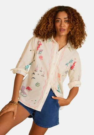 Kvinde med krøllet hår iført en knappet cremefarvet skjorte med farverige nautiske og blomsterprint, kombineret med blå denimshorts.