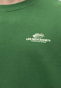 Πράσινο μπλουζάκι με λευκό λογότυπο «LES DEUX SOCIETY» και γραφική εικόνα υδρογείου στο στήθος, συνοδευόμενο από το κείμενο «PASSION FOR PEOPLE.»