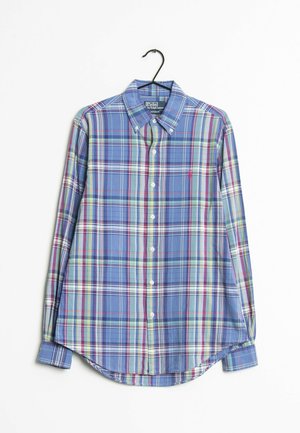 Chemise à manches longues à carreaux bleus, boutonnée avec col, suspendue sur un cintre noir sur fond blanc.