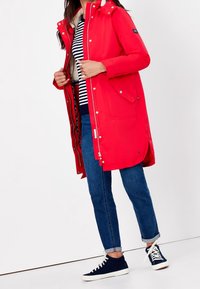 Parka impermeable roja con capucha y cierre de cremallera, con bolsillos laterales y un forro interior suave, combinada con zapatillas navy y jeans azules.