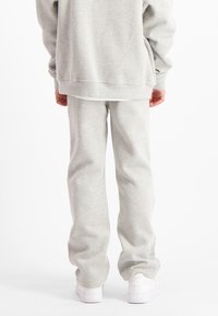 Persoon in lichtgrijze sweatpants en bijpassend sweatshirt, staand met de rug naar de camera, witte sneakers zichtbaar op een witte achtergrond.