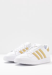 Pár bílých tenisek Adidas se zlatými pruhy a akcenty, bílými tkaničkami a texturovanou podrážkou na bílé ploše.