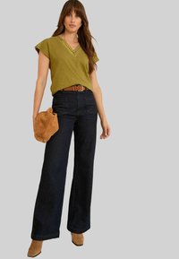 Moteris apsivilkusi alyvuogių žalios spalvos V formos iškirptės bliuską su akių detalėmis, tamsias plačias džinsines kelnes, rudą diržą, tan spalvos zomšinius batus ir laiko rudą zomšos rankinę.