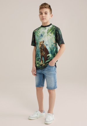T-shirt s krátkym rukávom s grafickým motívom tigra a džungľovými prvkami; kombinovaný s džínovými šortkami a bielymi teniskami s modrými akcentmi.