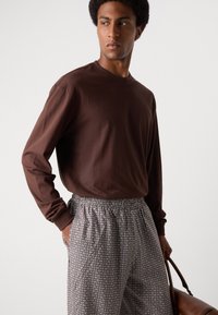 Chemise marron à manches longues associée à un pantalon gris à motifs avec une ceinture élastique et des poches, le tout accessoirisé avec un sac en cuir marron.