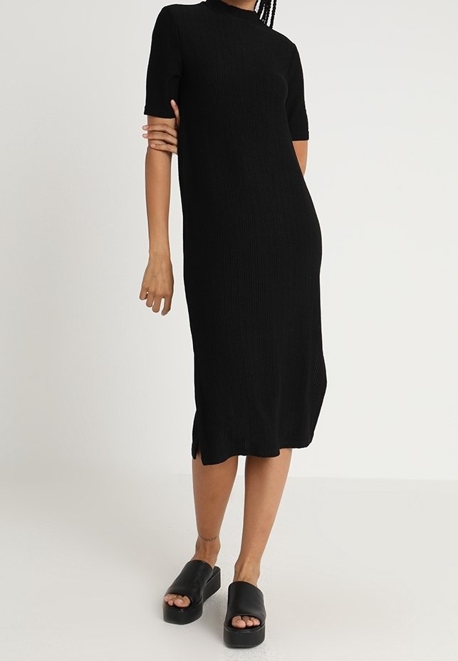 Femme portant une robe midi noire à manches courtes côtelée avec des fentes latérales et des sandales à plateforme noires, debout devant un fond uni.