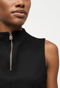 Prim-plan cu o persoană care poartă un top negru fără mâneci, cu un fermoar argintiu ce are inscripționat numele brandului „HUGO” pe elementul de prindere.