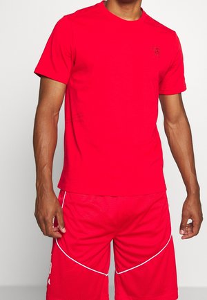 Hombre vestido con una camiseta de manga corta roja brillante y pantalones cortos deportivos rojos a juego con detalles de ribete blanco, de pie frente a un fondo sencillo.