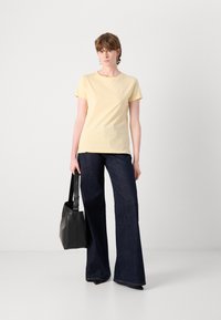 T-shirt gialla a maniche corte con scollatura rotonda, abbinata a jeans a campana blu scuro. Il modello tiene una borsa tote nera. Stile semplice e vestibilità casual.