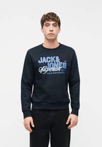 Νέος άνδρας που φοράει μαύρο φούτερ με την επιγραφή “JACK & JONES Superior New Aesthetics”, σε συνδυασμό με μαύρο παντελόνι, στέκεται μπροστά σε απλό φόντο.