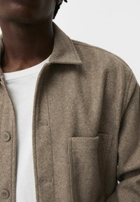 Braune Hemdjacke aus Wollmischgewebe mit Kragen, Knopfleiste und einer aufgesetzten Tasche. Sie zeichnet sich durch ein weiches, strukturiertes Gewebe und feine Nahtdetails aus.