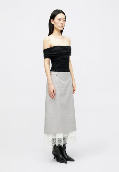 Moteris stovi vilkėdama juodą raukinį ofšoulder viršų, pilką midi sijoną su baltu nėriniu, juodus smailėjančius batus ir sidabro kaklo papuošalą.