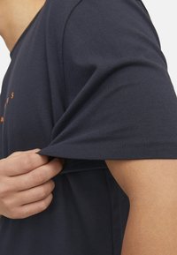 Maglietta di cotone blu navy con maniche corte, che mostra una mano che afferra la manica. Presenta un collo rotondo e nessun motivo o accessorio visibile.