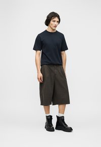 Νέος άνδρας που στέκεται φορώντας μαύρο μπλουζάκι, σκούρα φαρδιά σορτς μέχρι το γόνατο, λευκές κάλτσες και μαύρα δερμάτινα μποτάκια με κορδόνια.