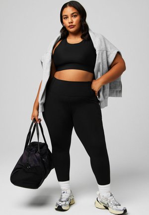 Frau in schwarzer Sportbekleidung mit grauem Sweatshirt über den Schultern, hält eine schwarze Sporttasche, trägt weiße Socken und silberne Turnschuhe vor einem schlichten Hintergrund.