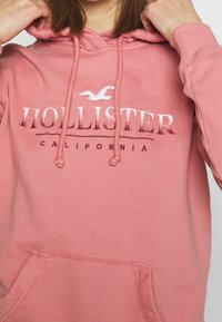 Rosa huvtröja med dragsnören, framficka och texten "HOLLISTER CALIFORNIA". Mjukt tyg med en slät textur och logodetalj.