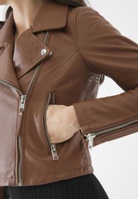 Giubbotto da motociclista in pelle marrone con cerniere e accessori in argento. Presenta colli inclinati, tasche laterali e un design aderente. Finitura testurizzata.